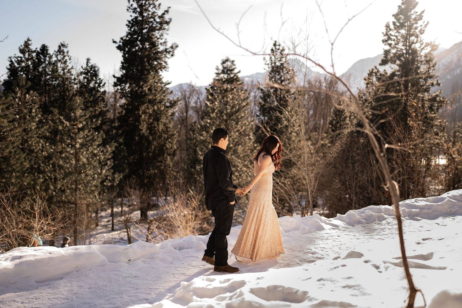 A couple stands together in winter sunlight with mountains and snow-covered trees behind them.