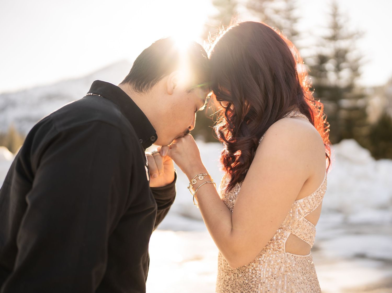 A backlit couple shares an intimate moment in the snow with golden sunlight filtering through.