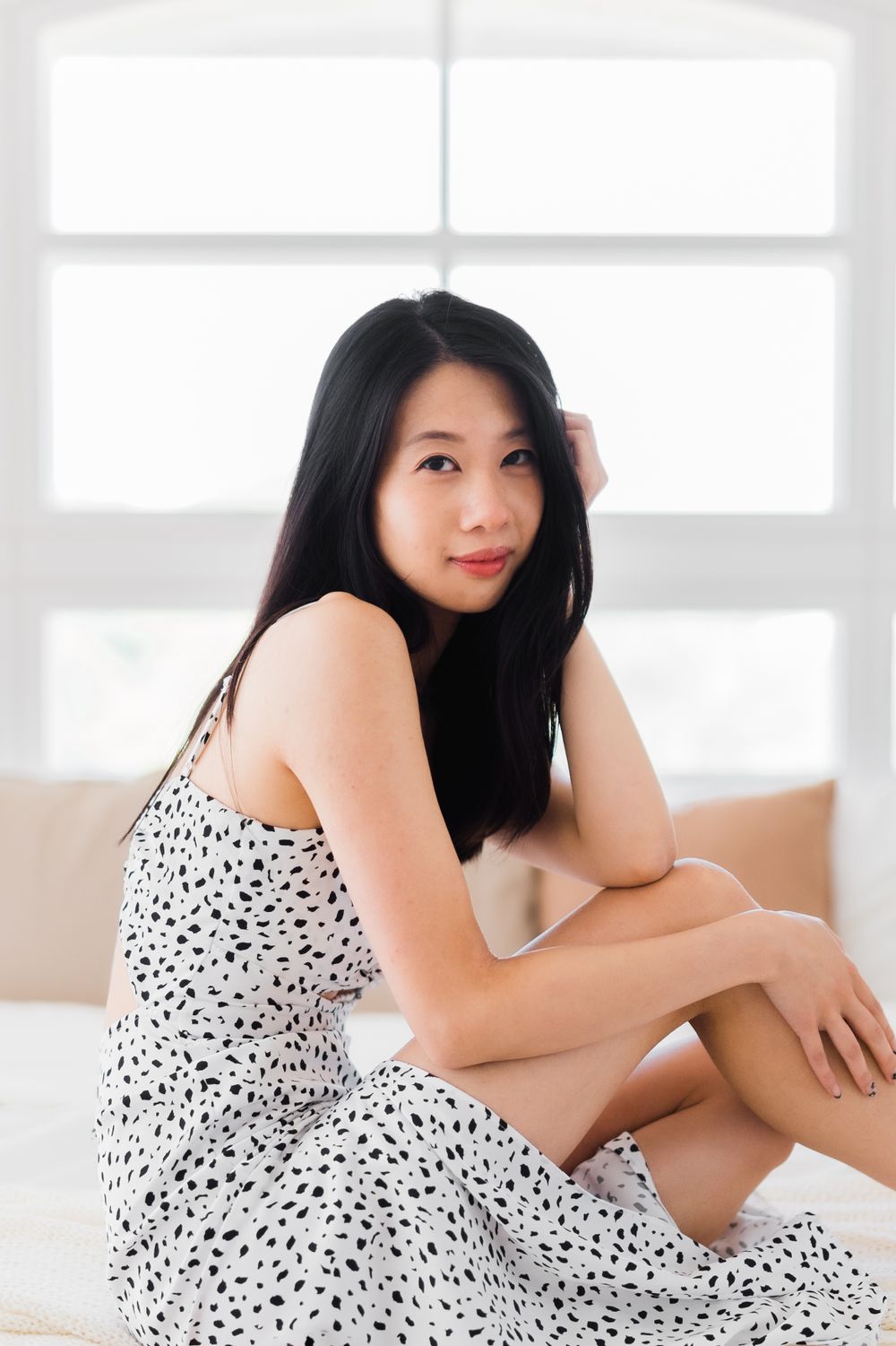 Figure in dotted dress poses elegantly on white bedding in sunlit room.