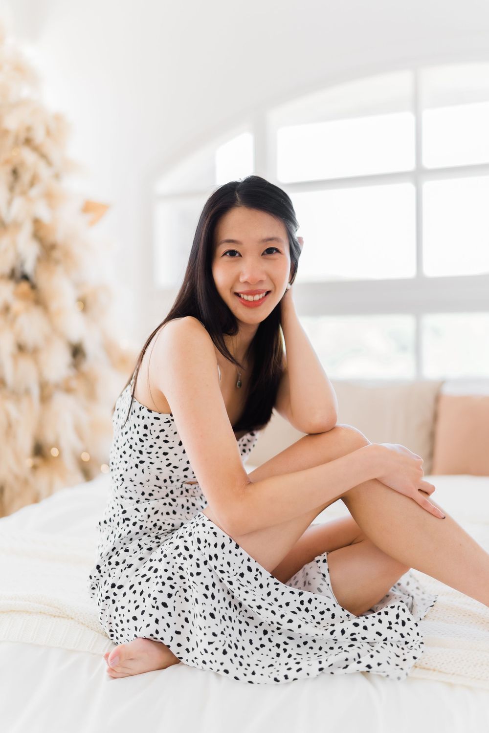 Person in spotted dress sits relaxed on white bed with natural lighting.