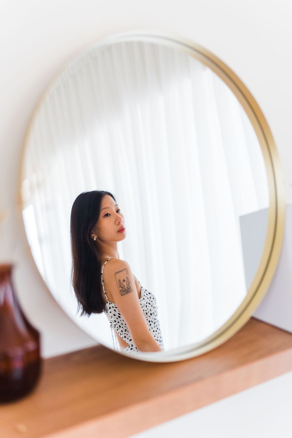 A round mirror captures a profile reflection with a spotted dress and tattoo visible.