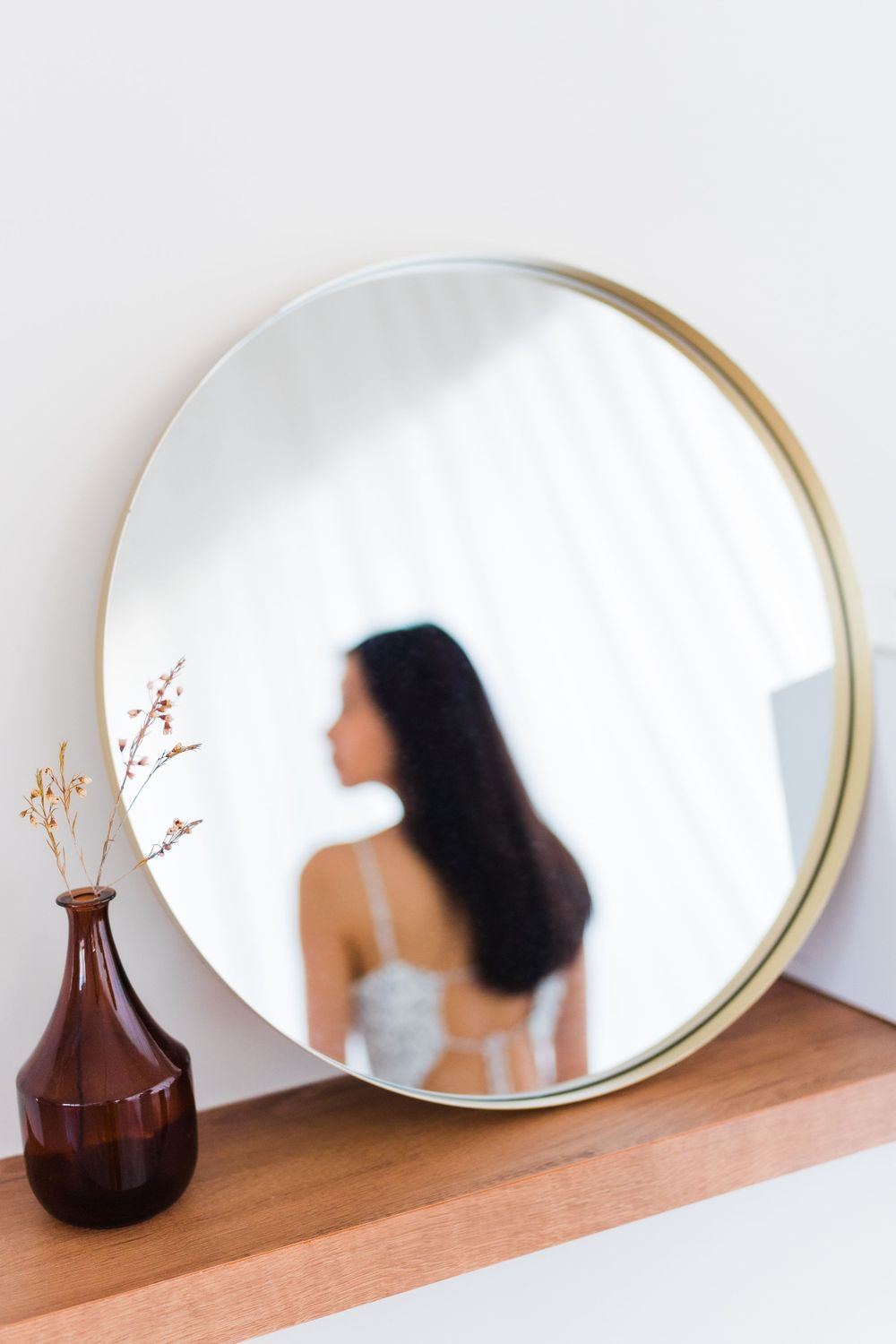 A circular gold mirror reflects a silhouette with long dark hair next to a brown vase.
