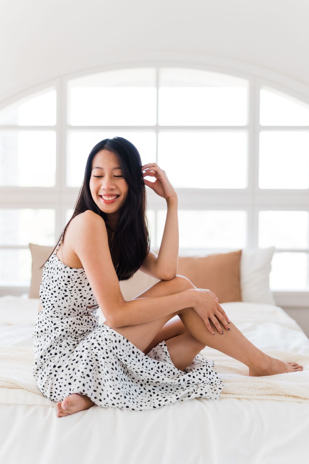 Person wearing spotted sundress sits casually on white bedding near bright window.
