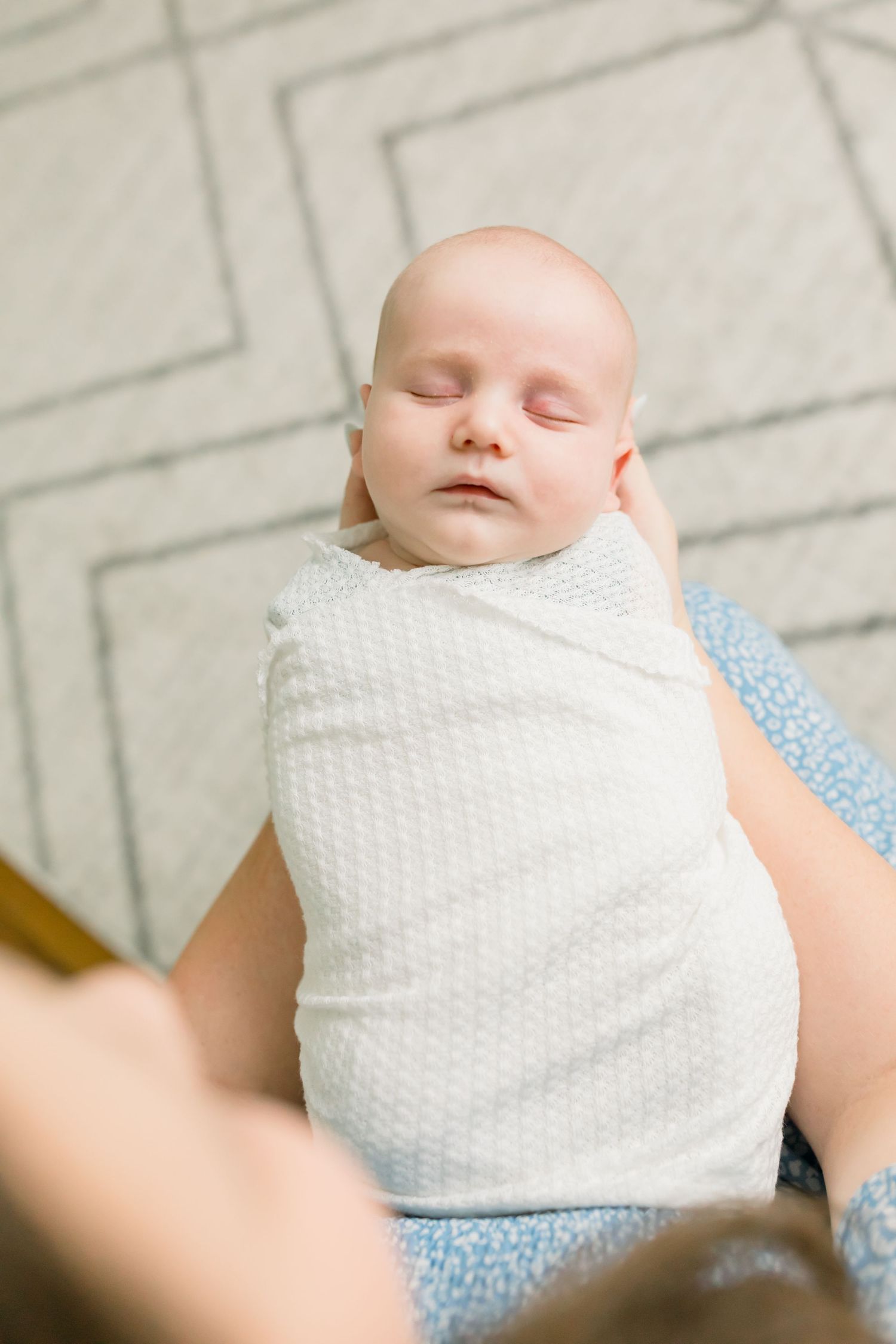 A sleeping baby wrapped in white swaddle on a geometric patterned rug.