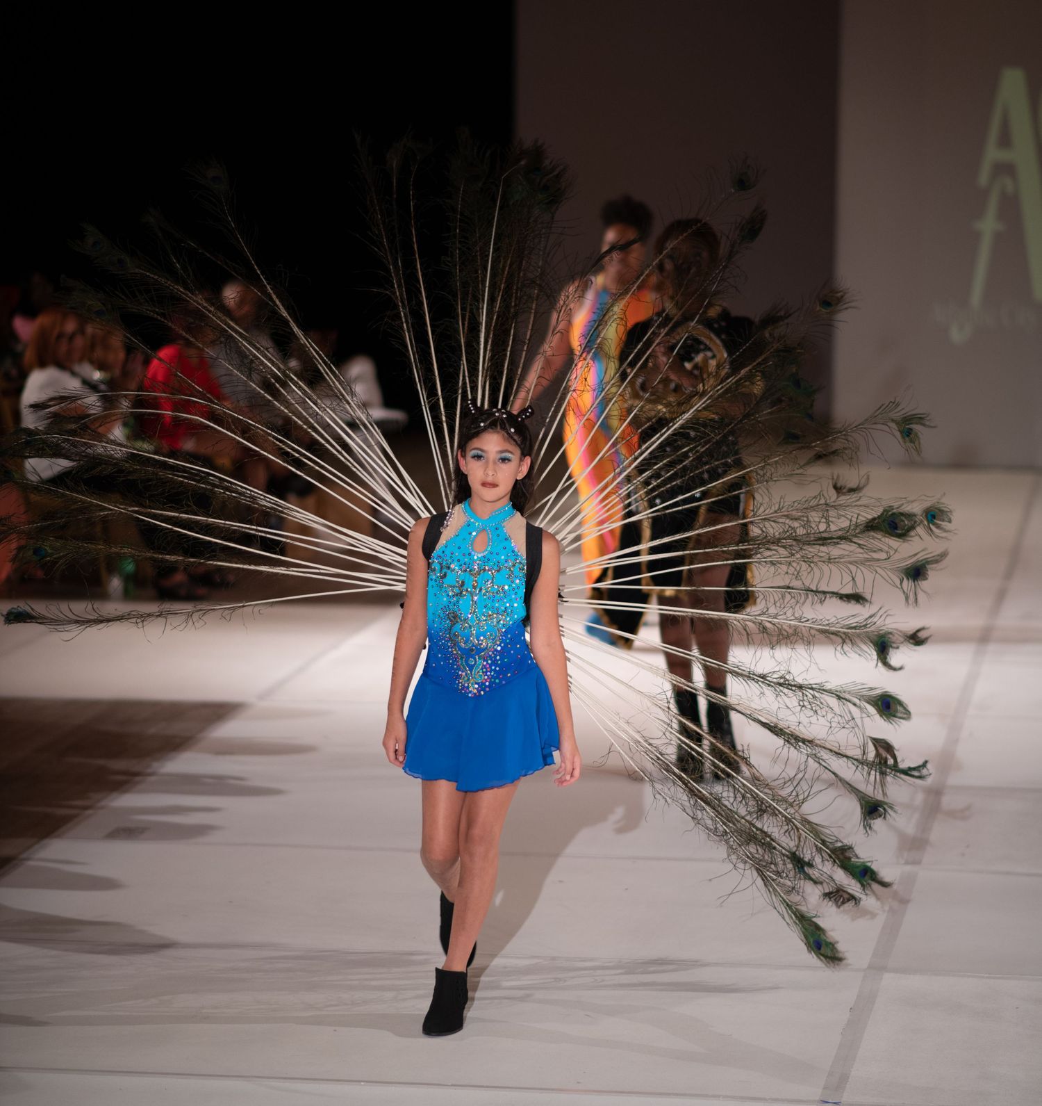 A model walks the runway in a blue dress with dramatic peacock feather wings.