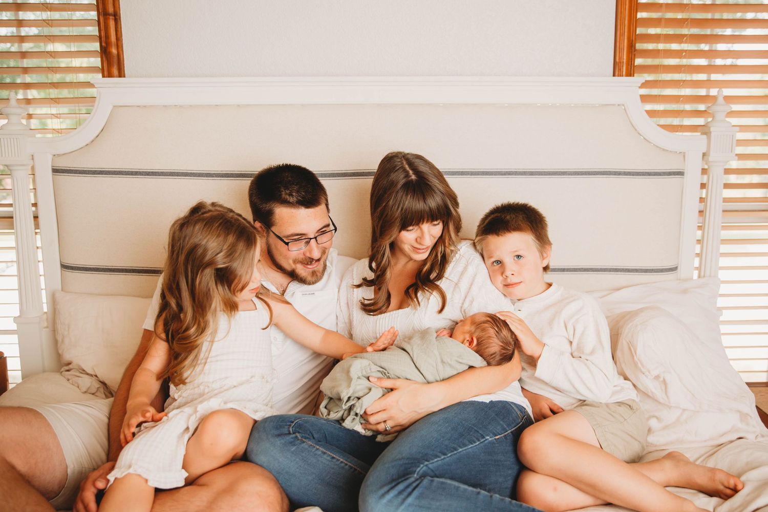 Family sitting together on white bed with newborn baby in casual indoor setting.