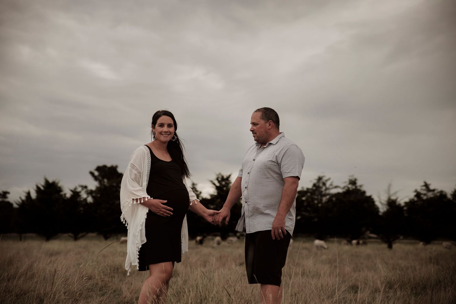 A couple holding hands stands together in a grassy field under moody overcast skies during a maternity photoshoot.