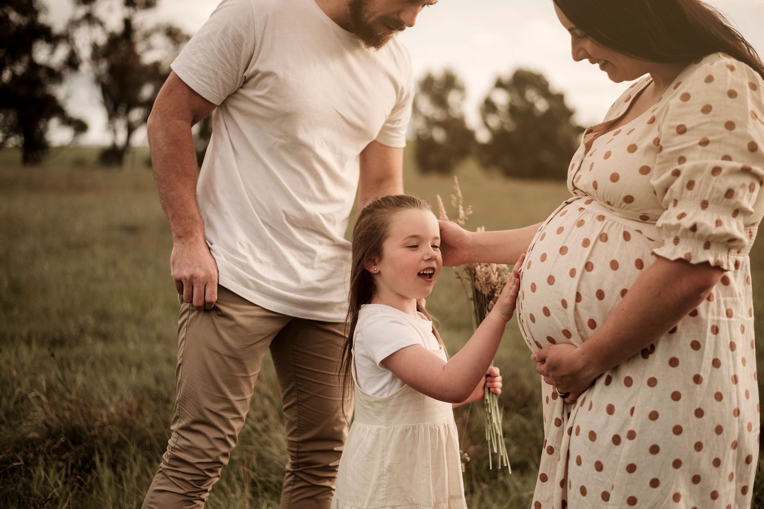 Family stands together in a field at sunset with young child touching pregnant mother's belly in polka dot dress.