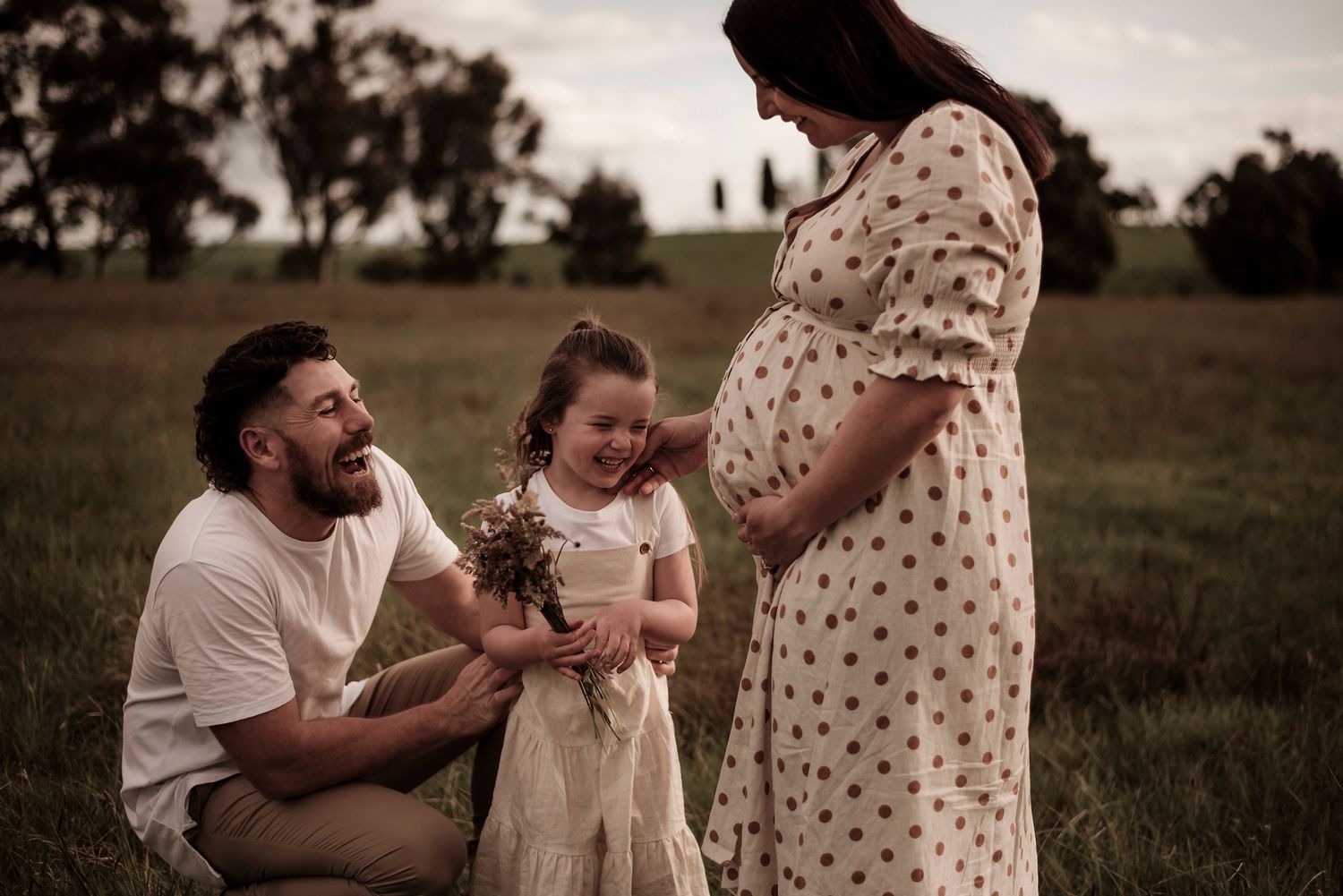 Expectant couple and young girl share a tender moment in a grassy field at sunset. Traralgon Railway Reserve.