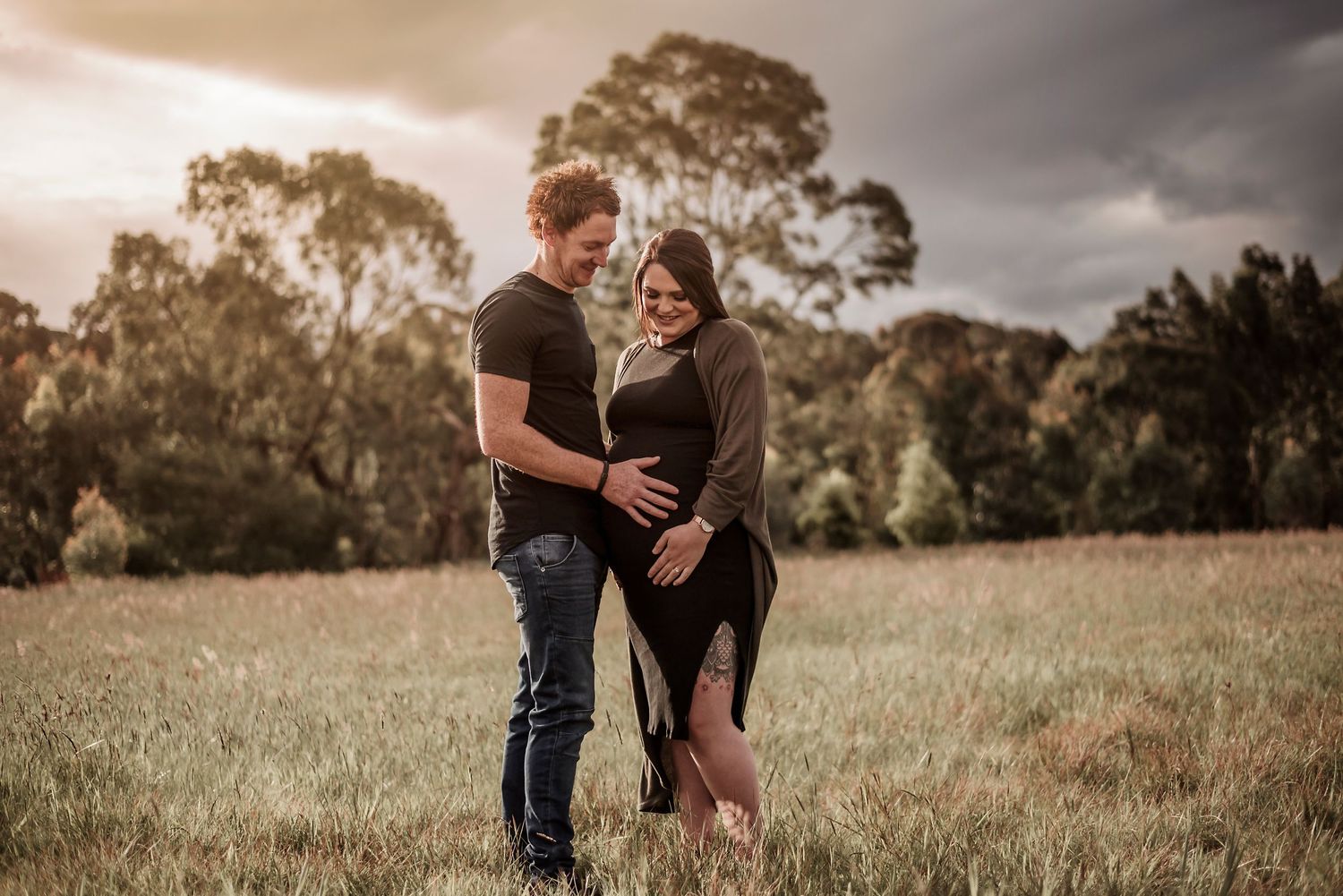 Romantic golden hour portrait of a couple embracing in a field with trees silhouetted against stormy skies.
