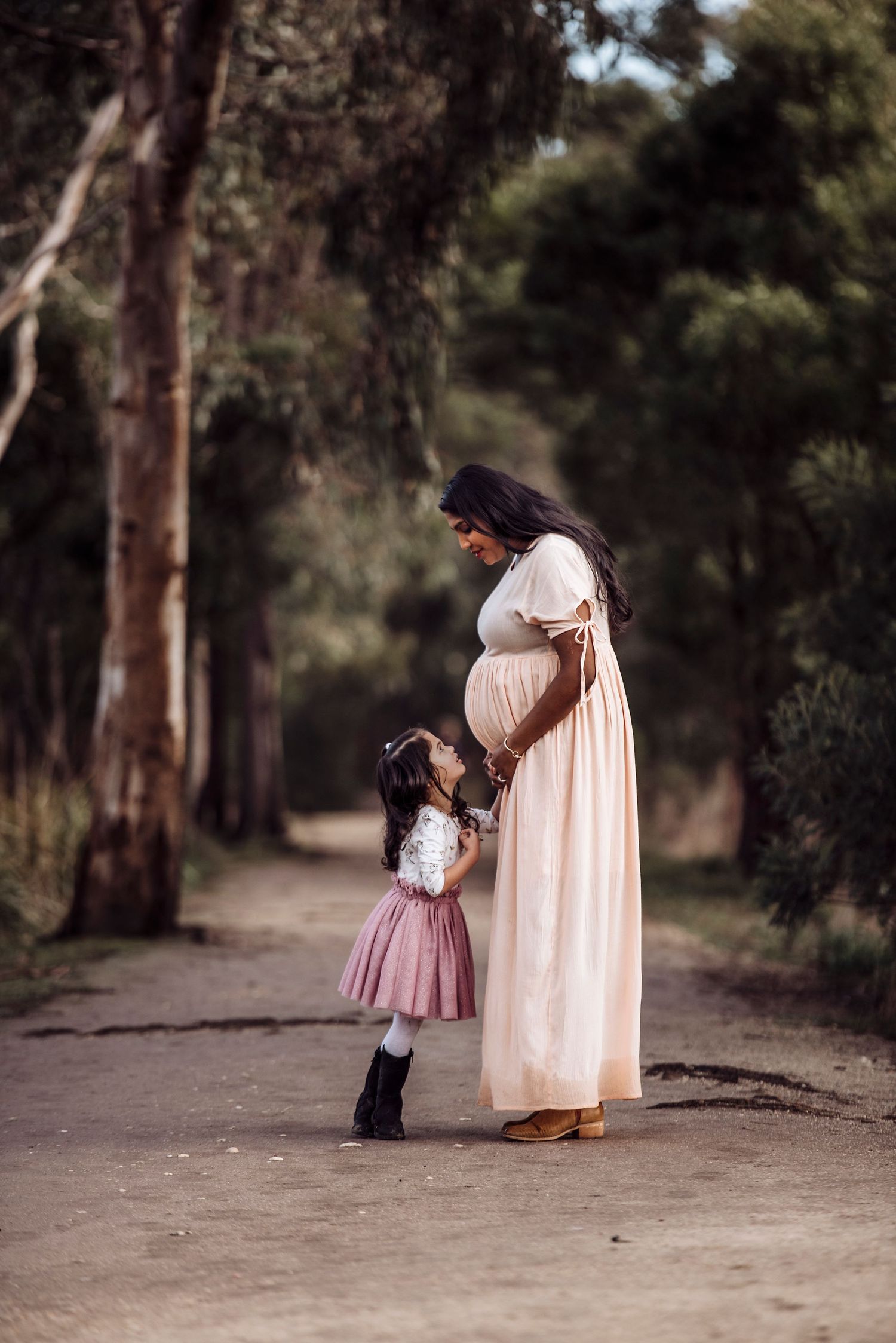 A tender moment between mother and child on a wooded path as they look at her pregnant belly.