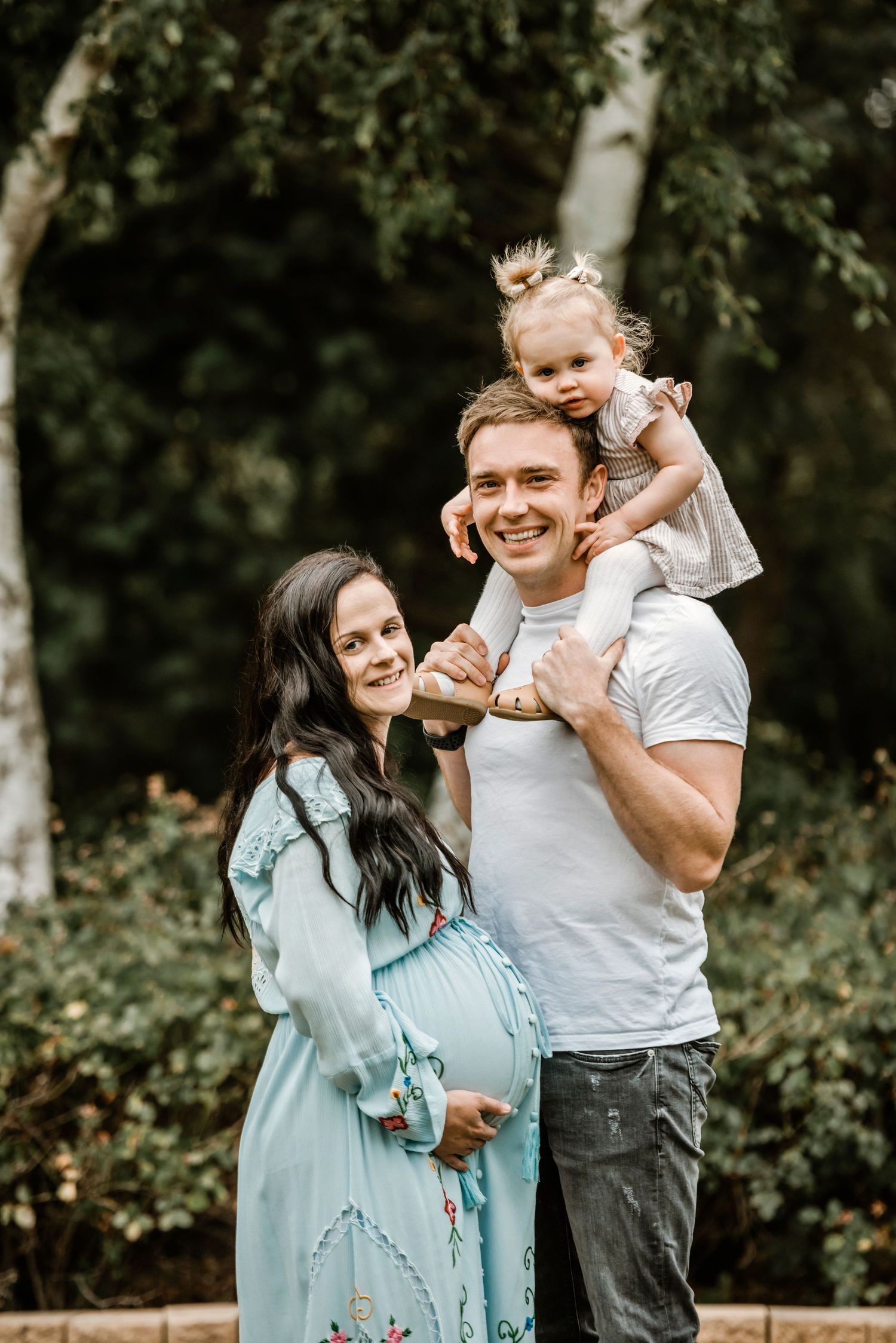 Expecting parents pose outdoors with their toddler while mother shows her baby bump in a blue dress.