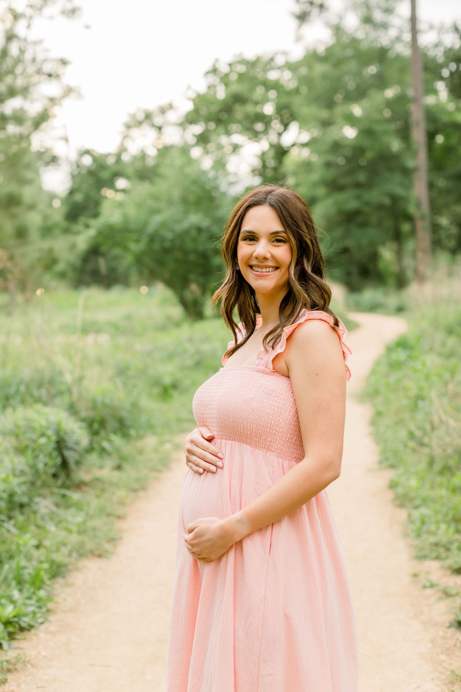 A woman in a pink empire waist dress smiles while cradling her baby bump on a garden path.