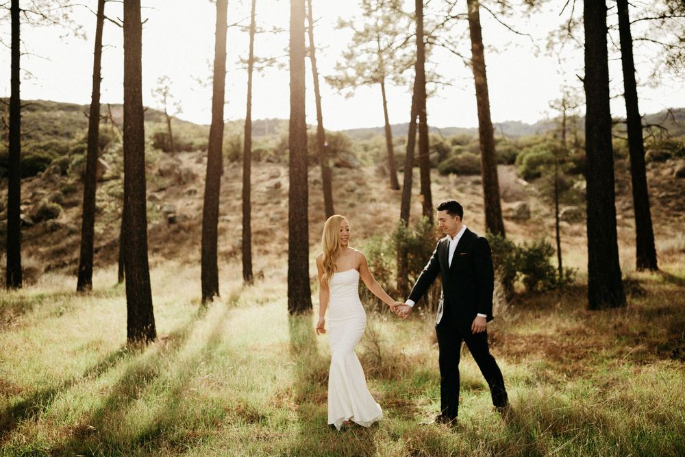 A couple in wedding attire hold hands while walking through a sunlit forest clearing.