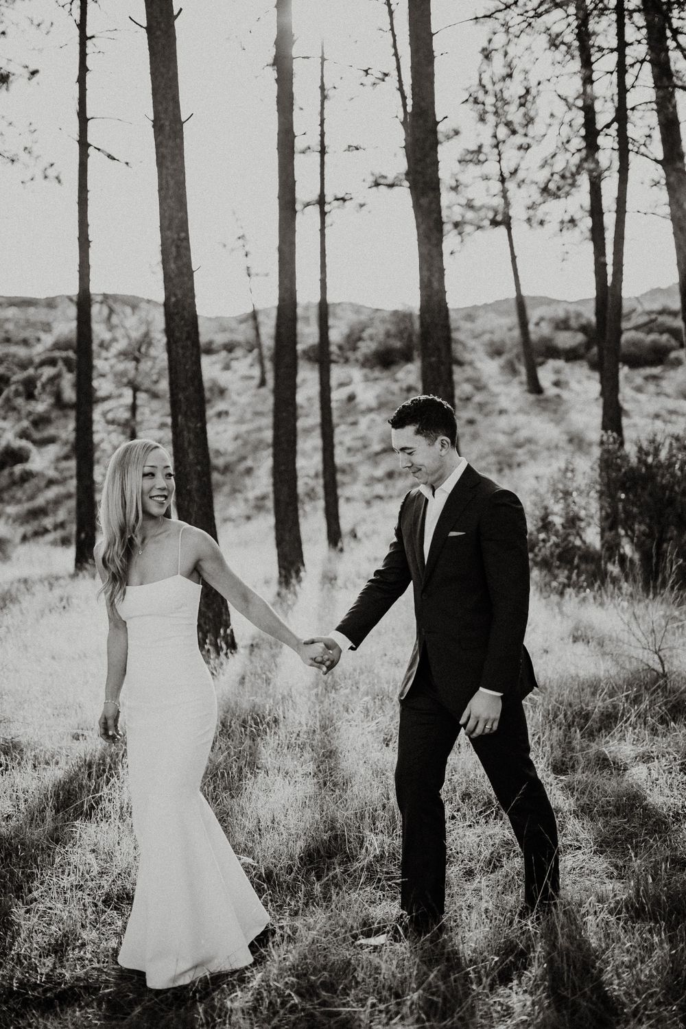 Black and white photo of a couple in wedding attire walking through a pine forest.