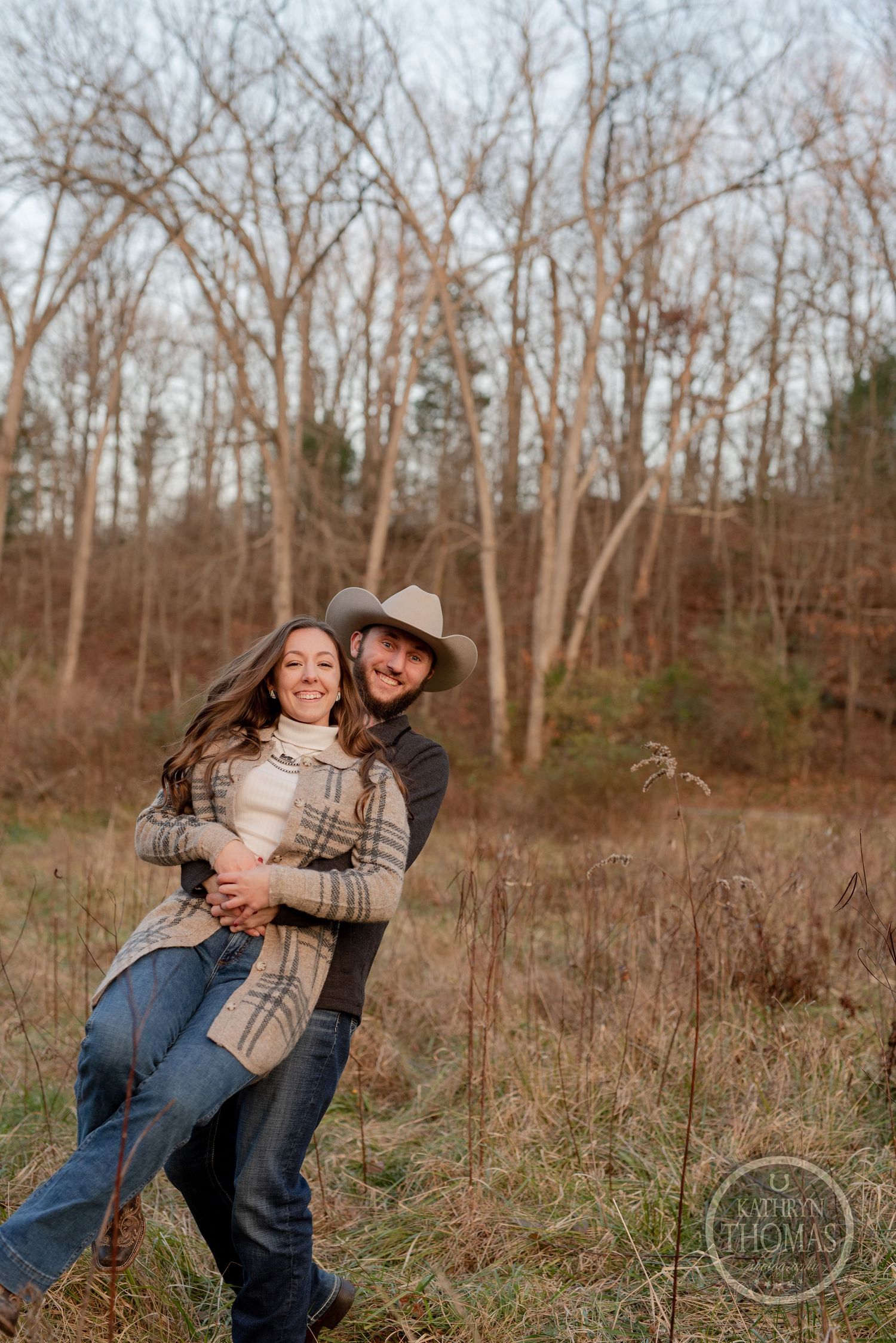 A joyful embrace captured in a rustic field with bare trees in the background.