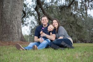 Mom and Dad sitting in the grass under a tree holding their baby.
