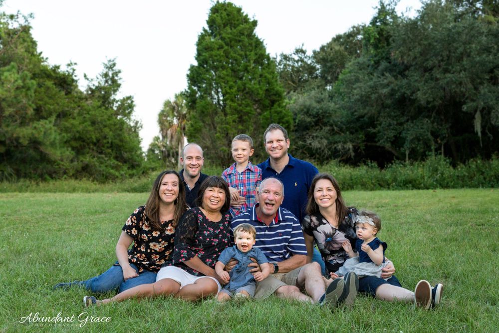Large family smiling and posing while sitting in the grass in a big field.