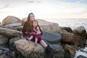 Mother holding her daughter on her lap while sitting on a large rock on the beach at sunset.