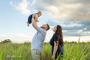 Dad holding his new baby in the air while mom and dad make faces at the baby standing a grassy field.