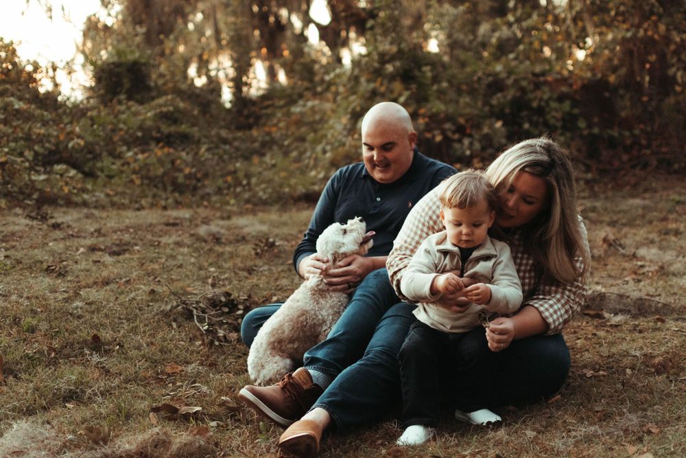 A family sits together outdoors on the ground during golden hour in a wooded setting.