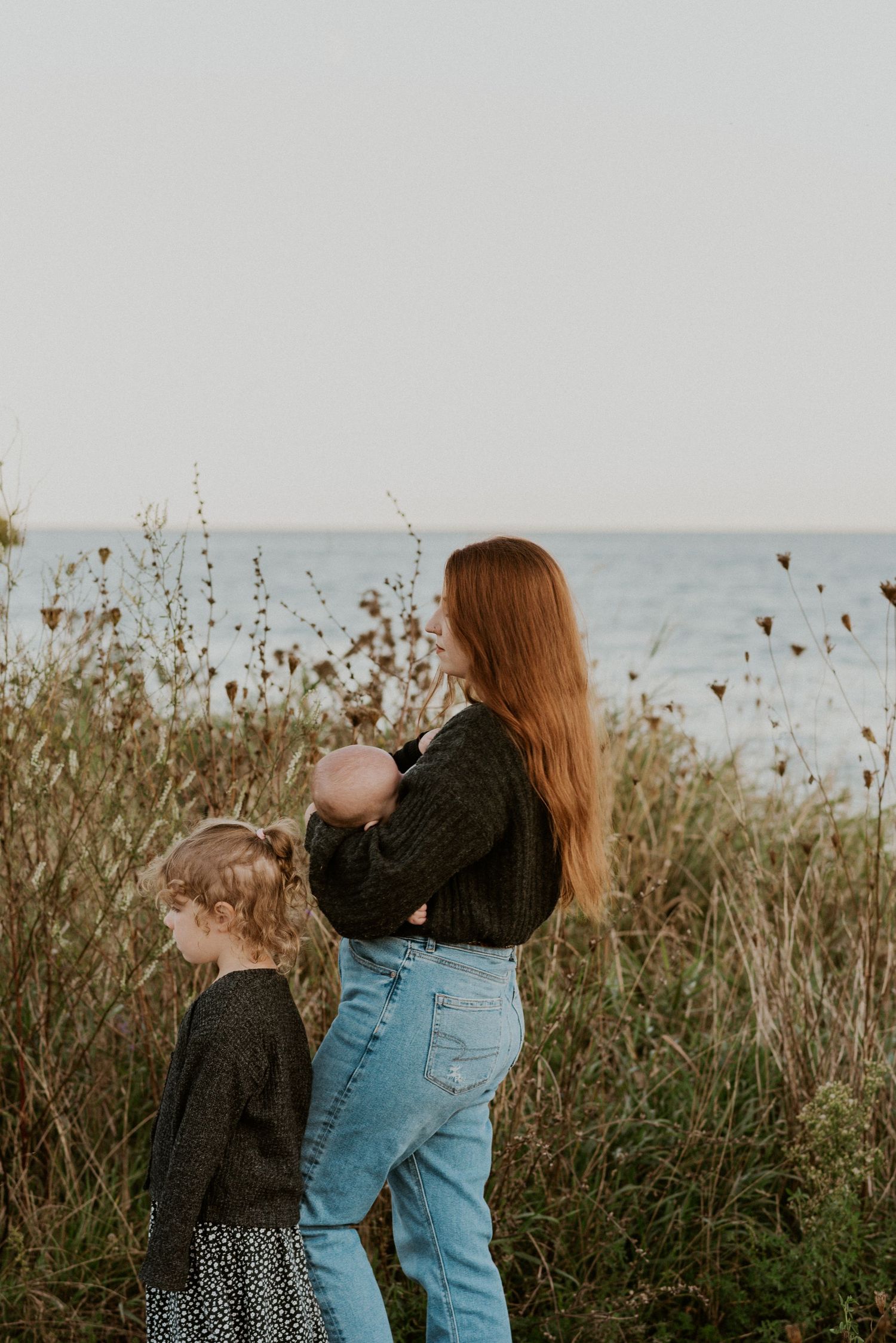 Mother and children standing among wildflowers at ocean's edge during golden hour.