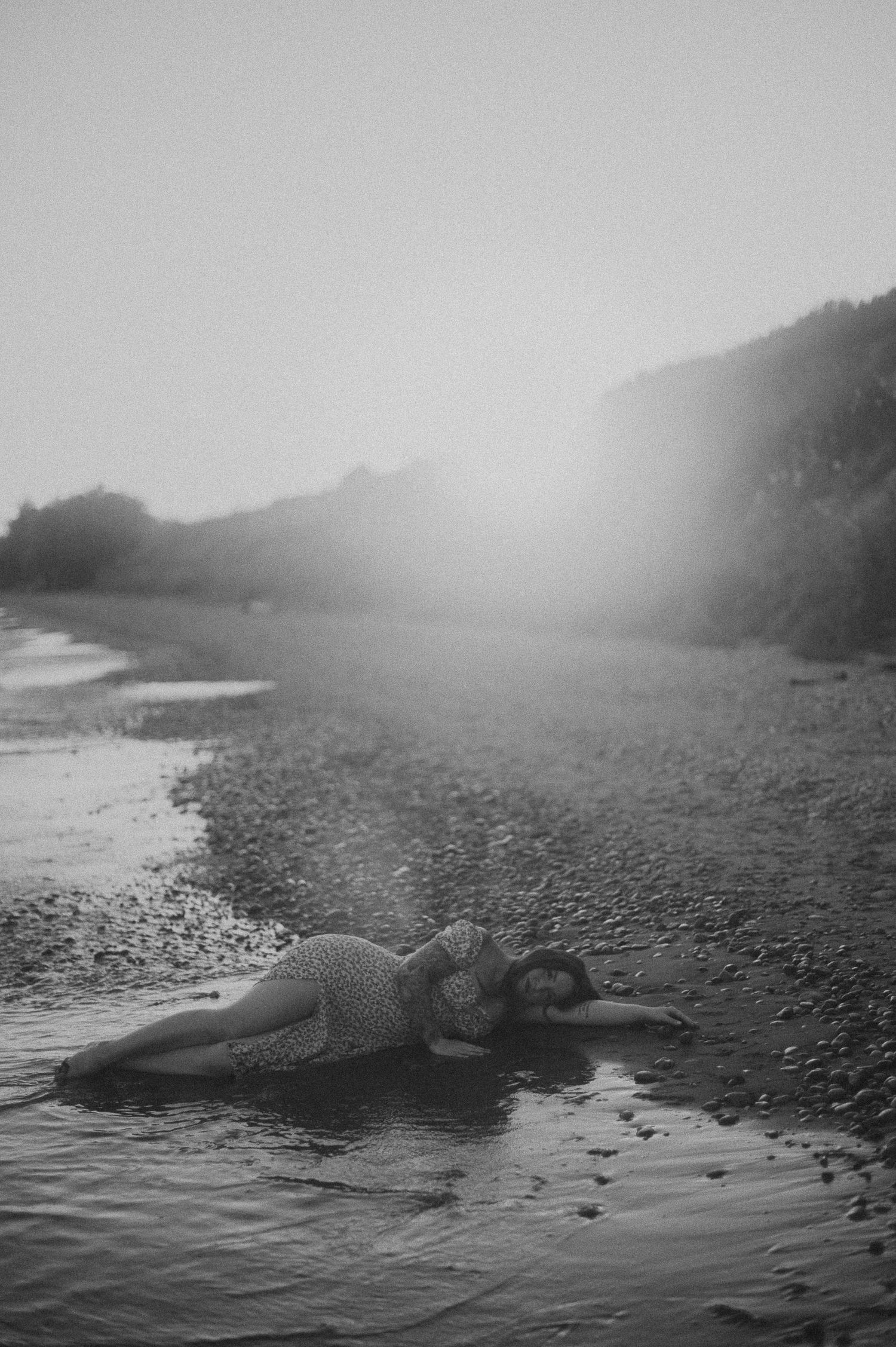 Moody black and white photo of someone lying on a misty beach at the water's edge.