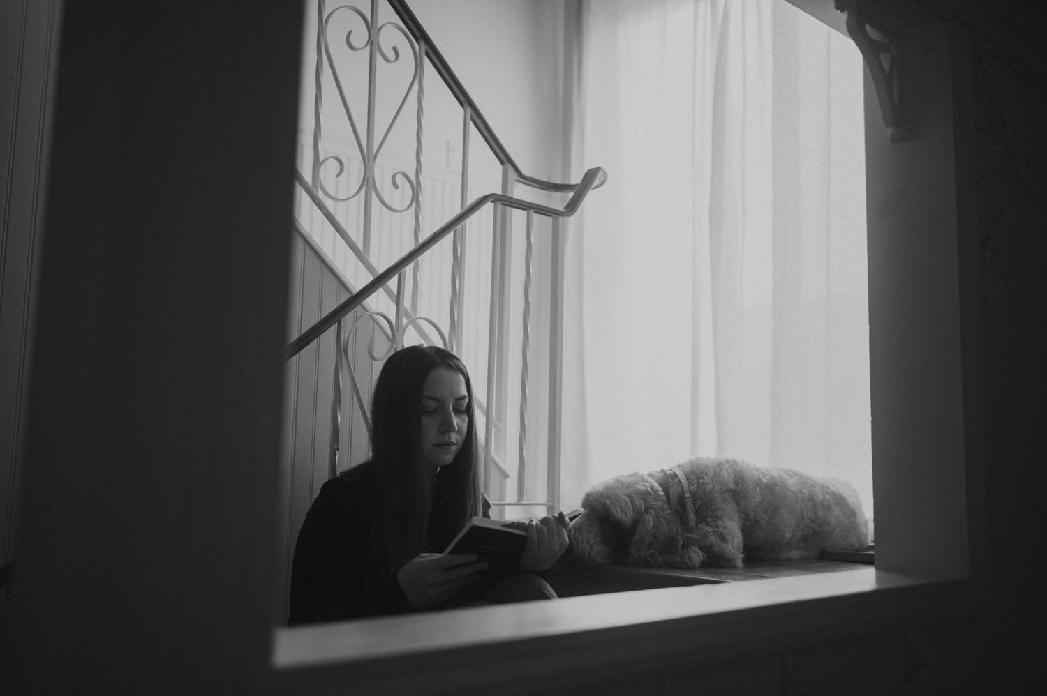Moody black and white image of person and cat resting by window with iron railing.