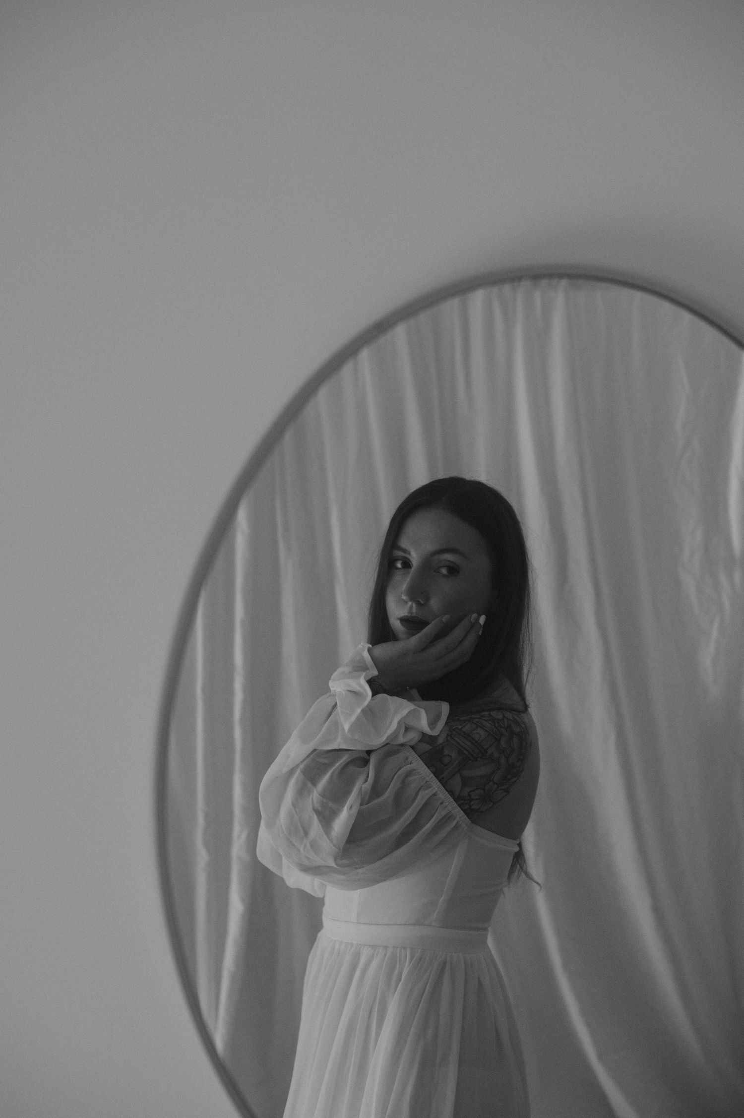 Artistic black and white portrait through a round mirror with white curtain backdrop.