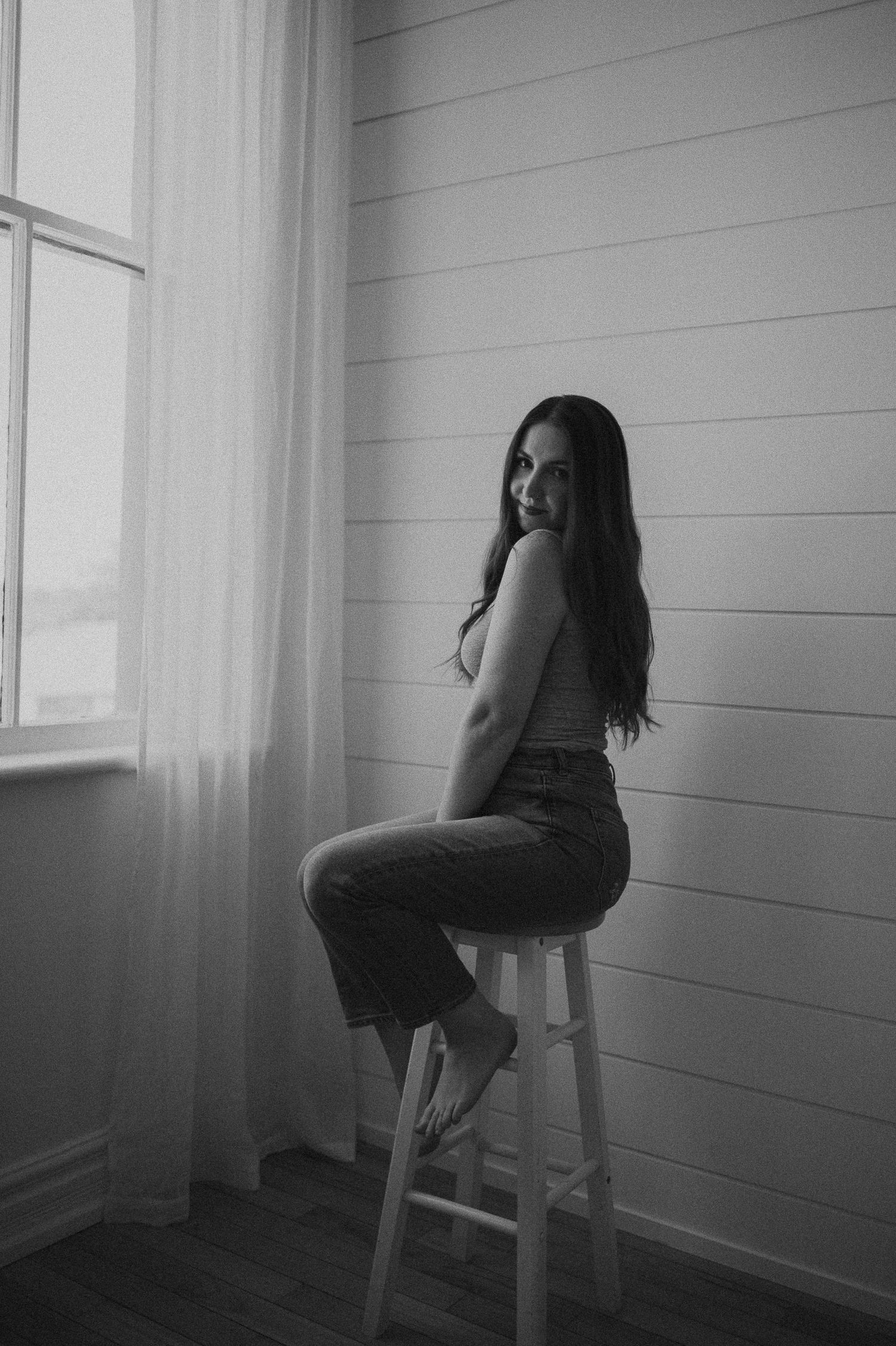 A black and white portrait of a woman sitting on a stool next to a window in a minimal white room.