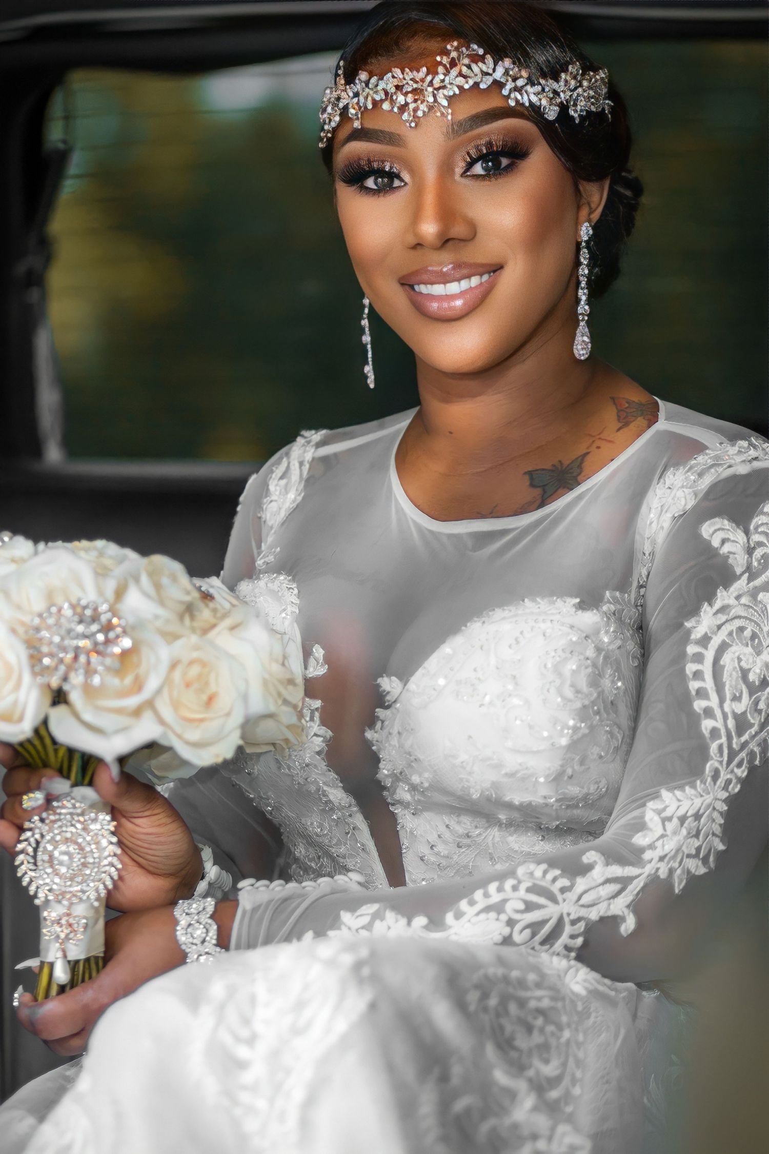 Elegant bride in lace long-sleeve gown holding white rose bouquet with crystal headpiece inside a vehicle.