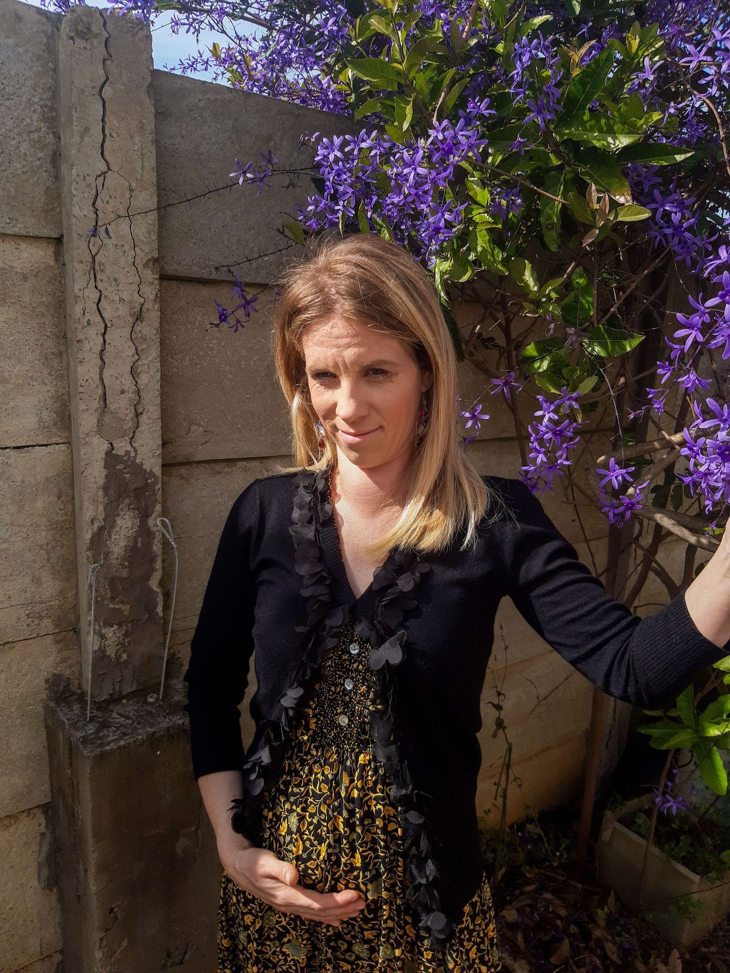 Person in black cardigan and floral dress posing beside purple wisteria blooms.