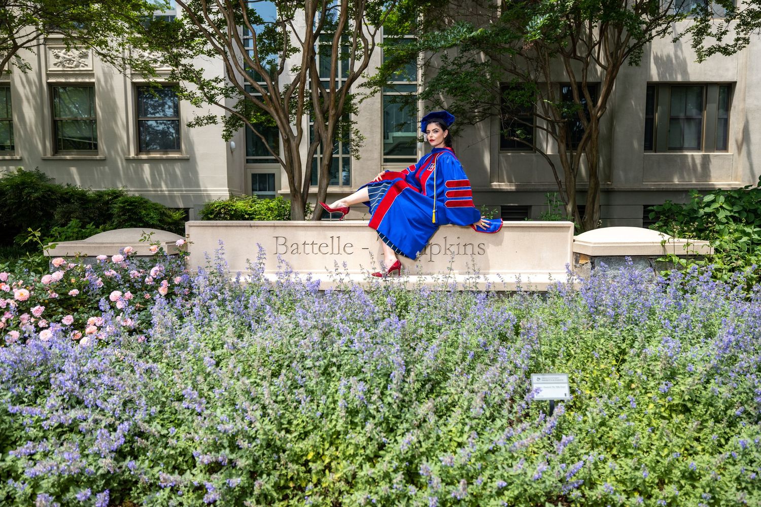 Graduate in academic regalia sits among blooming purple flowers on campus.