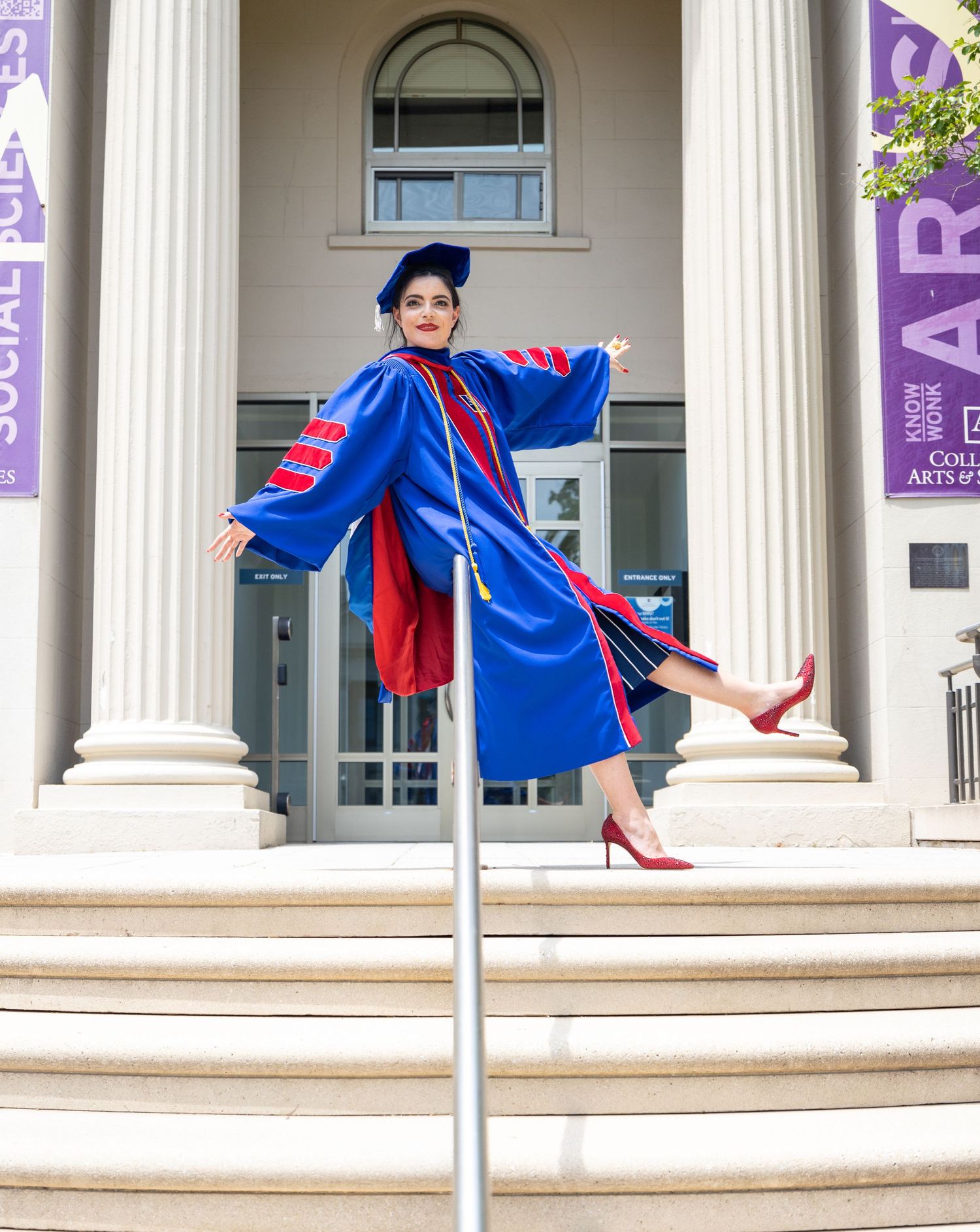 Graduate in blue regalia poses playfully on white stone steps between classical columns.
