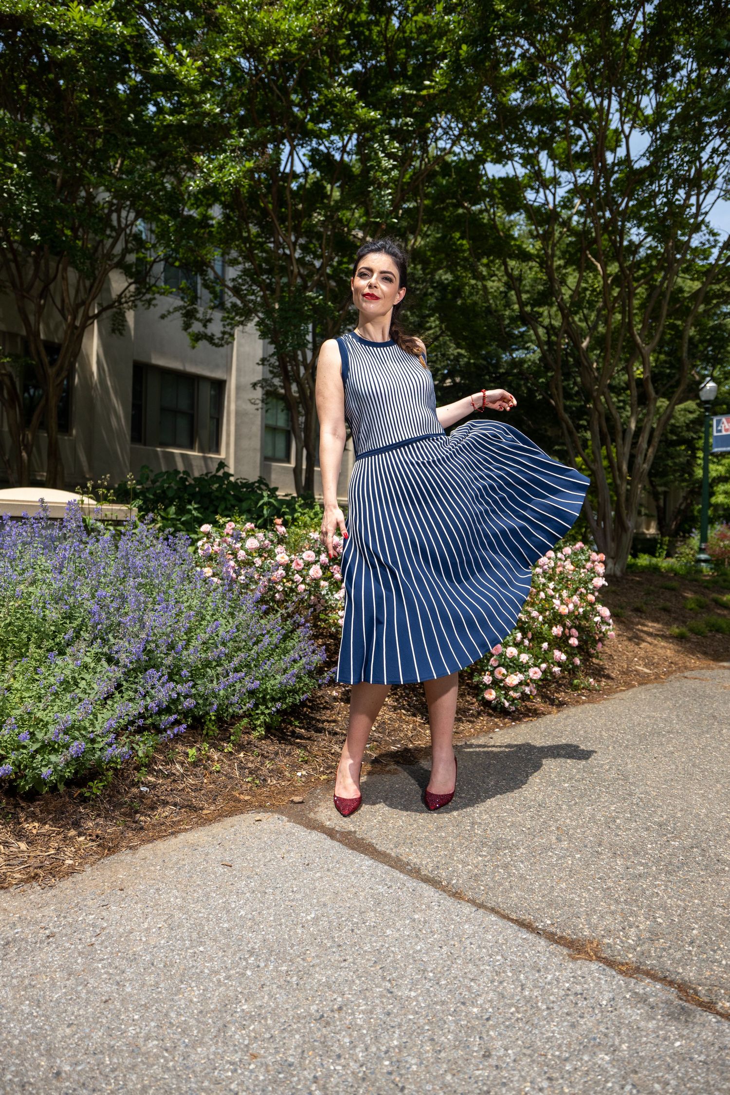 Woman twirling in navy pleated dress poses along garden pathway with blooming flowers.