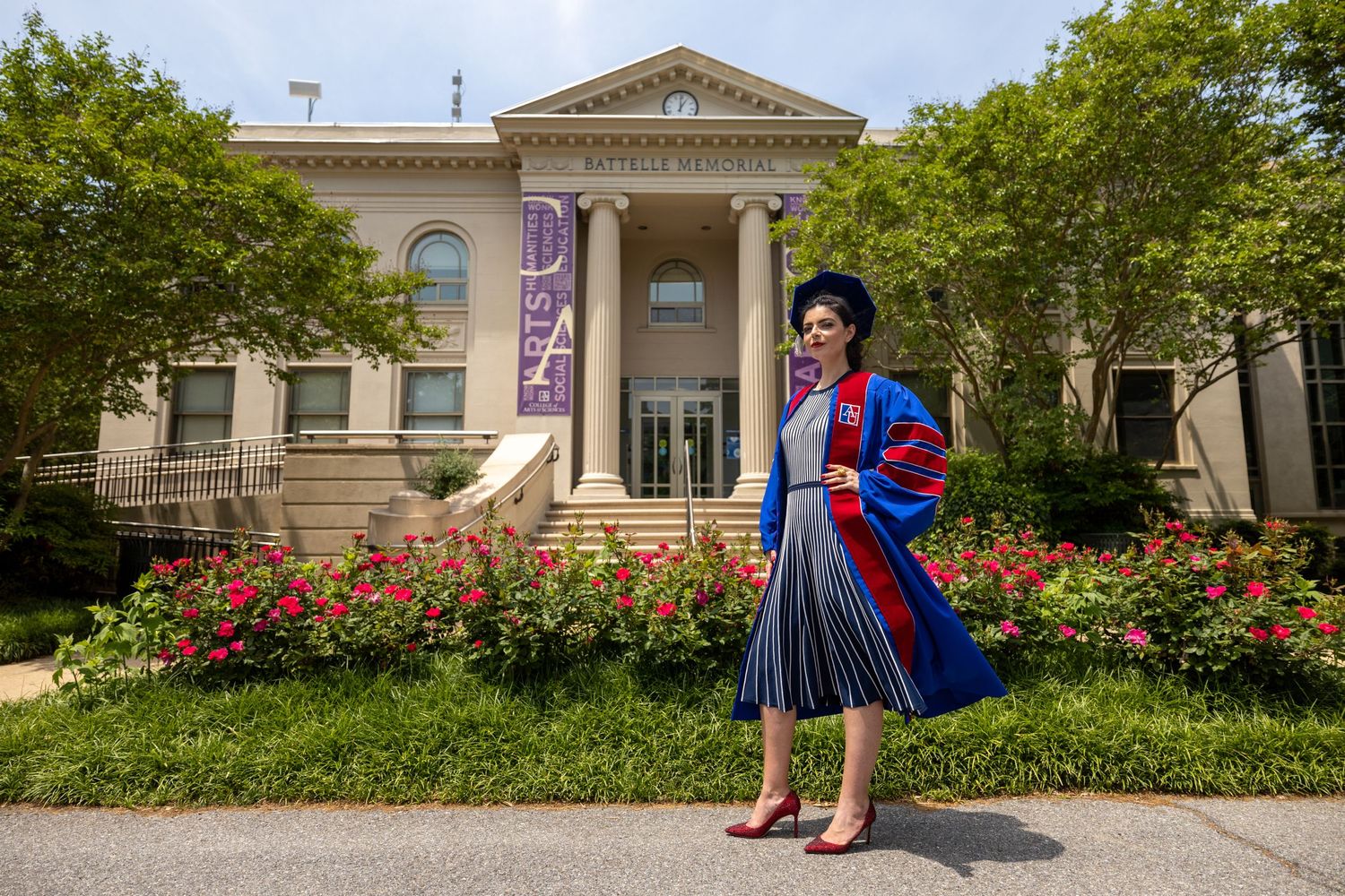 Graduate in academic regalia poses beside rose garden at classical university building.