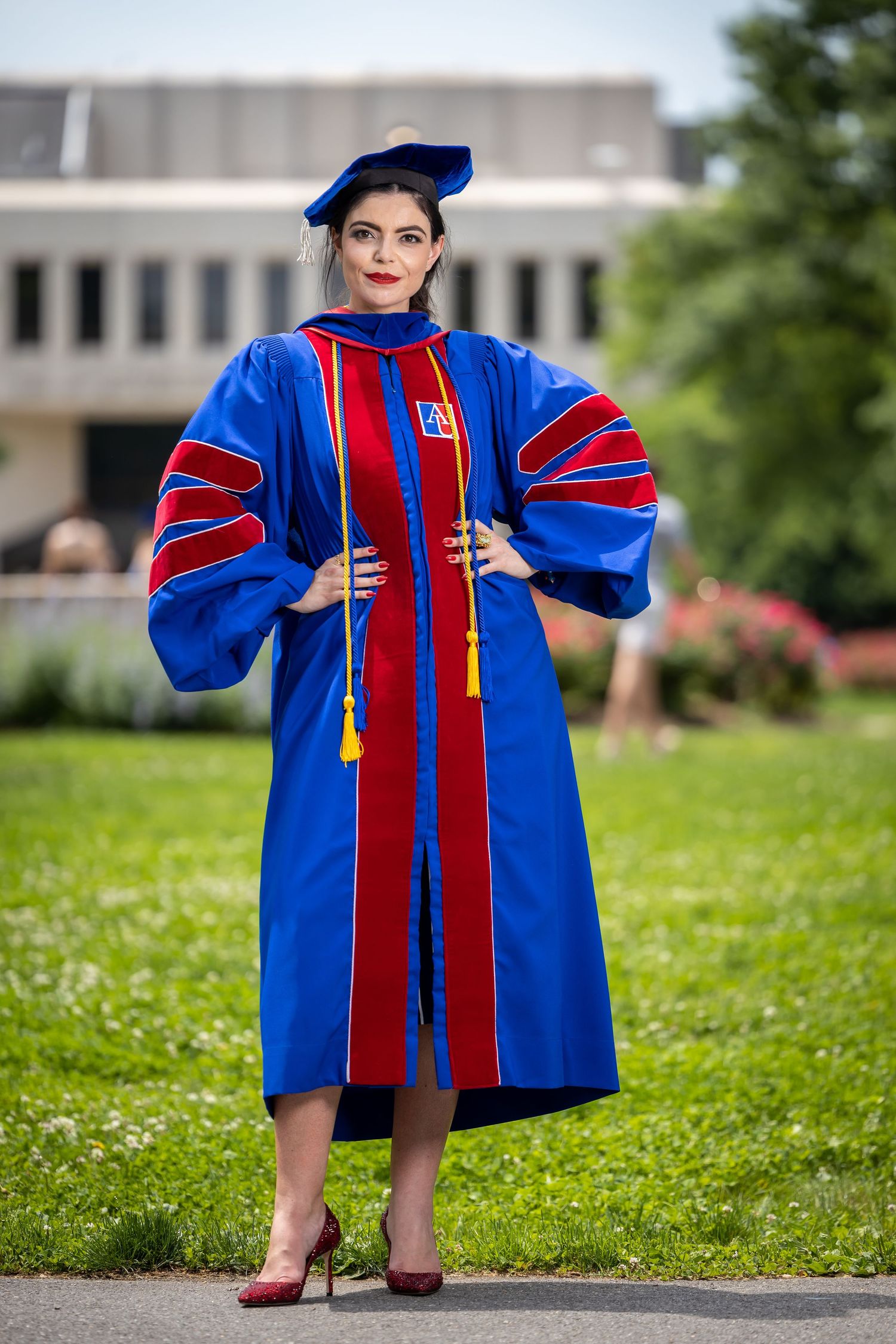 Doctoral graduate in blue and red academic regalia poses confidently on university grounds.