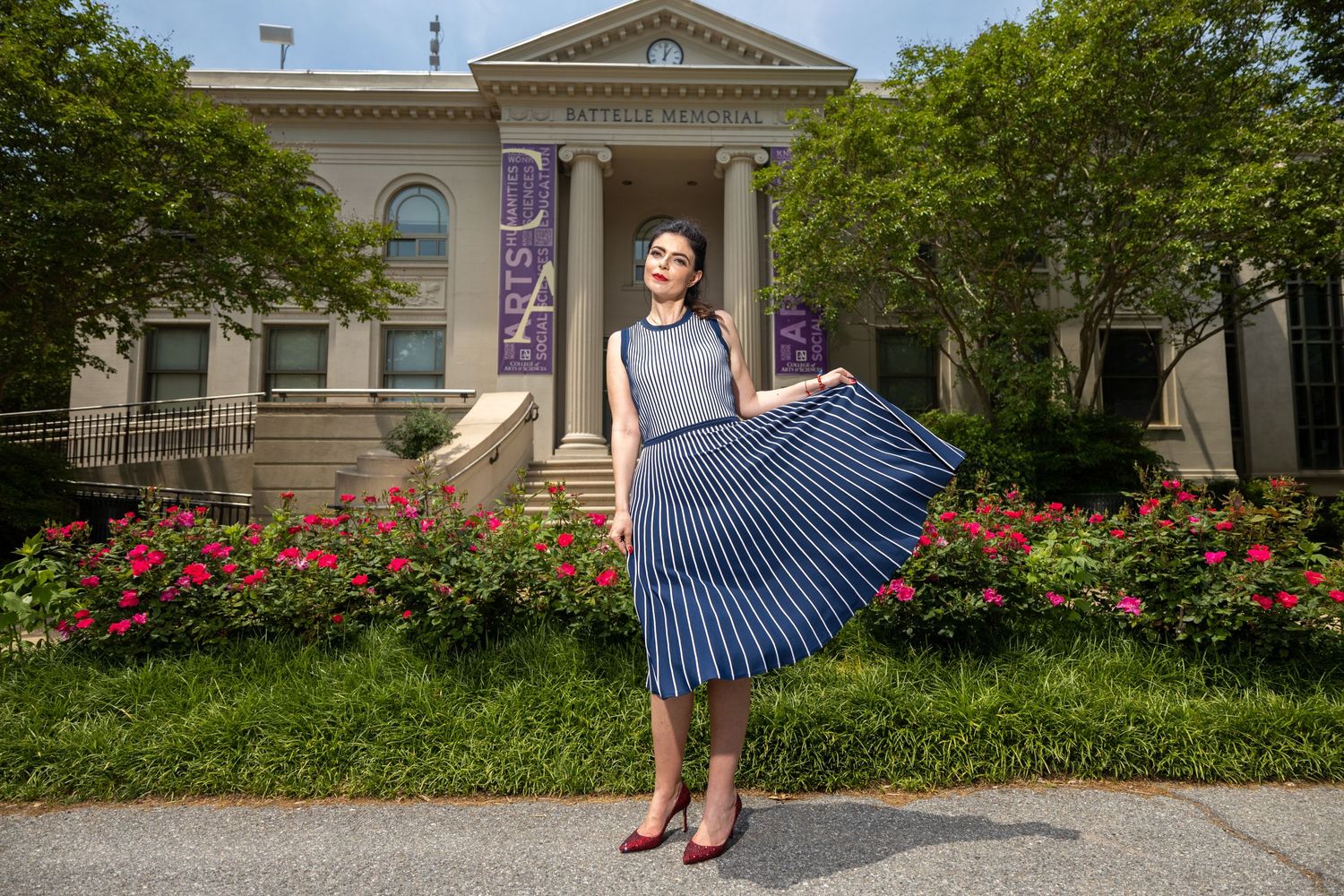 Woman in vintage-style blue striped dress poses gracefully in front of historic building with blooming flowers.