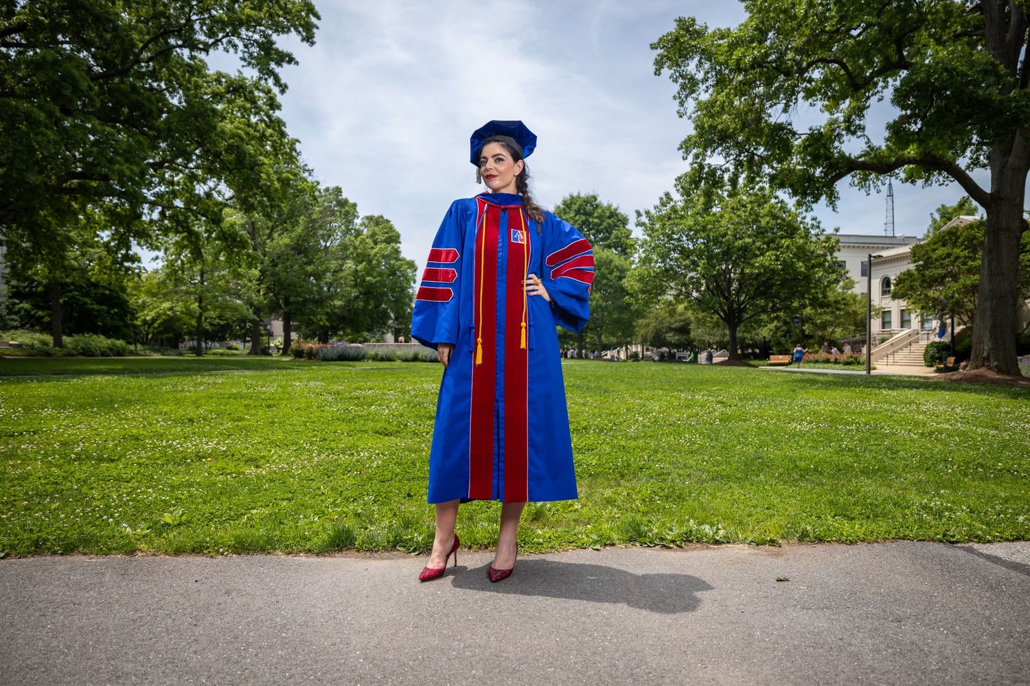 Graduate in traditional blue and red doctoral regalia stands on campus pathway.