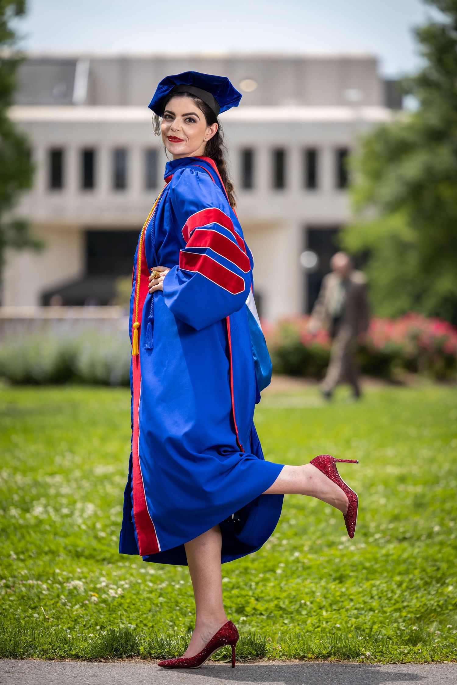 Graduate in ceremonial blue gown strikes playful pose on campus lawn.