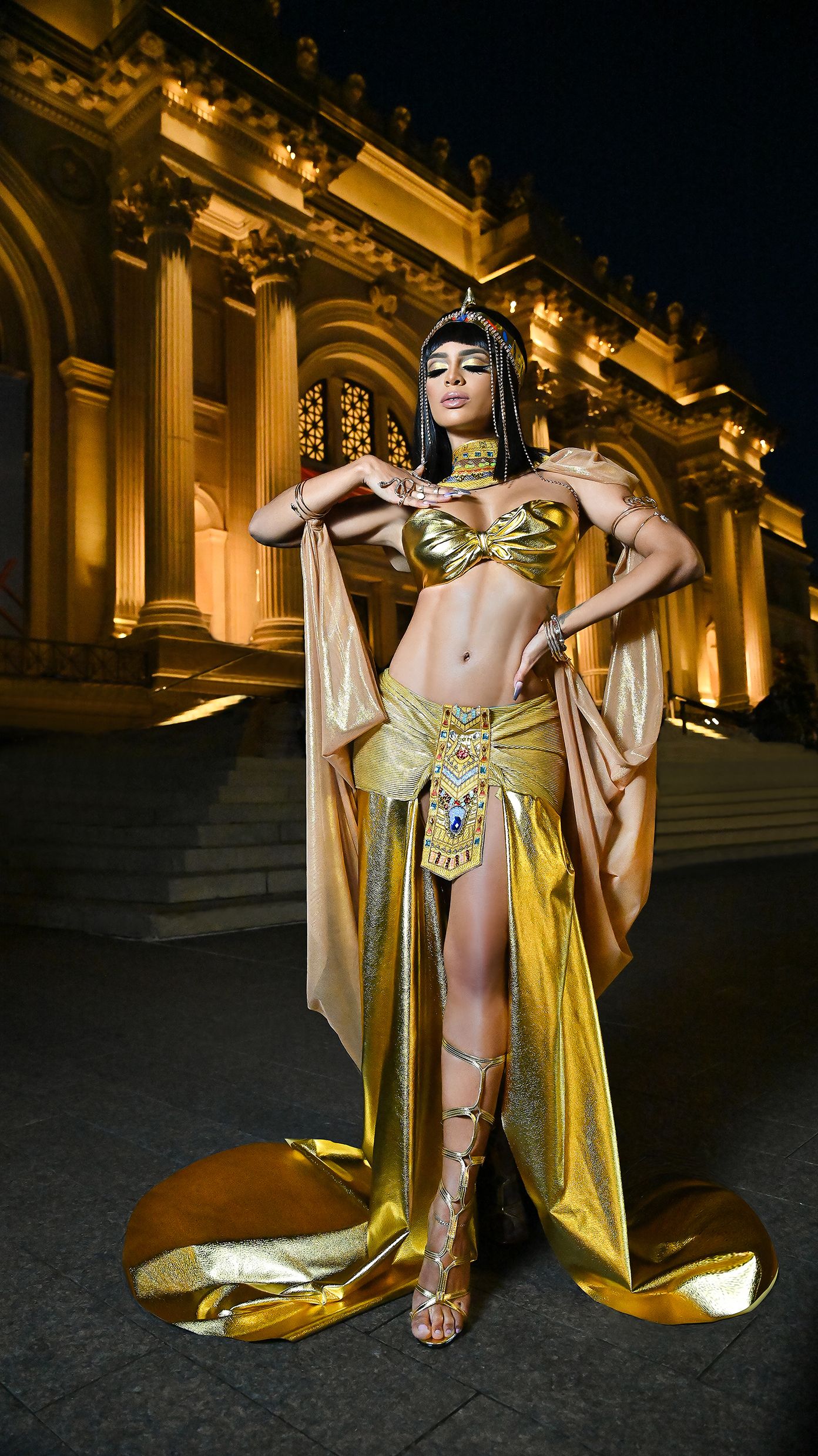 Egyptian-inspired costume with gold details and flowing cape photographed at night by classical building.