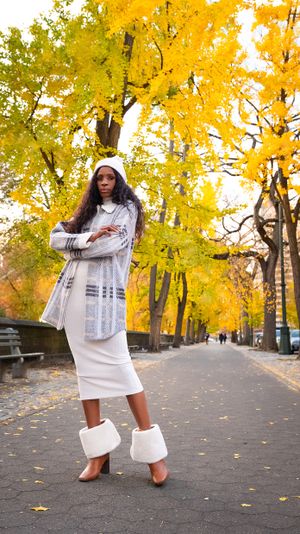 Fashionable autumn outfit featuring white dress, patterned cardigan and boots against yellow fall foliage.