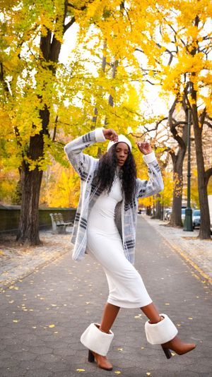 Dancing pose in white winter outfit with boots against golden autumn trees in park.