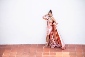 Glamorous pink satin evening gown with dramatic cape photographed against white wall.