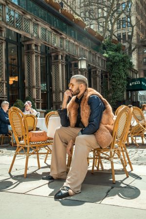 Relaxed pose at outdoor cafe featuring fur vest and casual wear against urban backdrop.