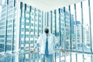 Person in white coat looking out large windows of modern office building with city view.