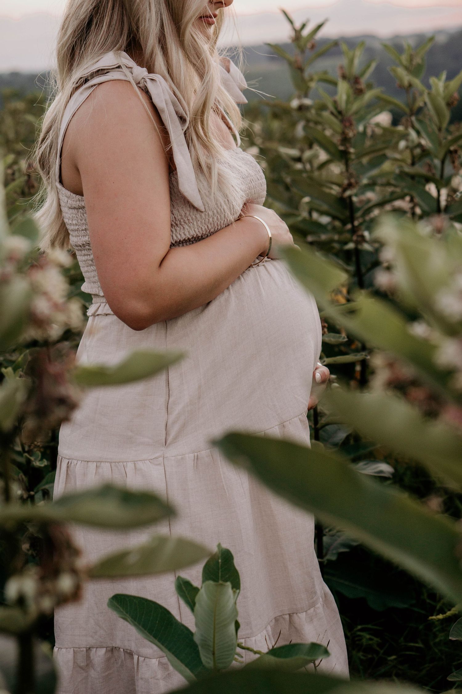 Pregnant woman in beige dress cradling baby bump while standing among blooming garden plants.