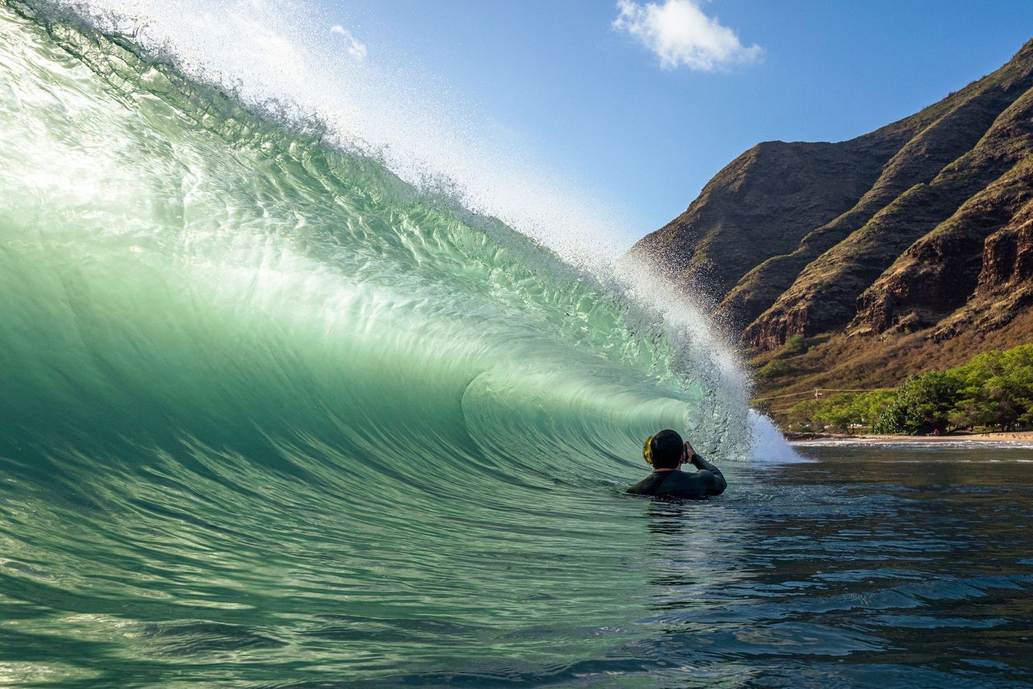 Emerald wave curls through crystal clear water with dramatic mountain cliffs in the background of a Hawaiian paradise.