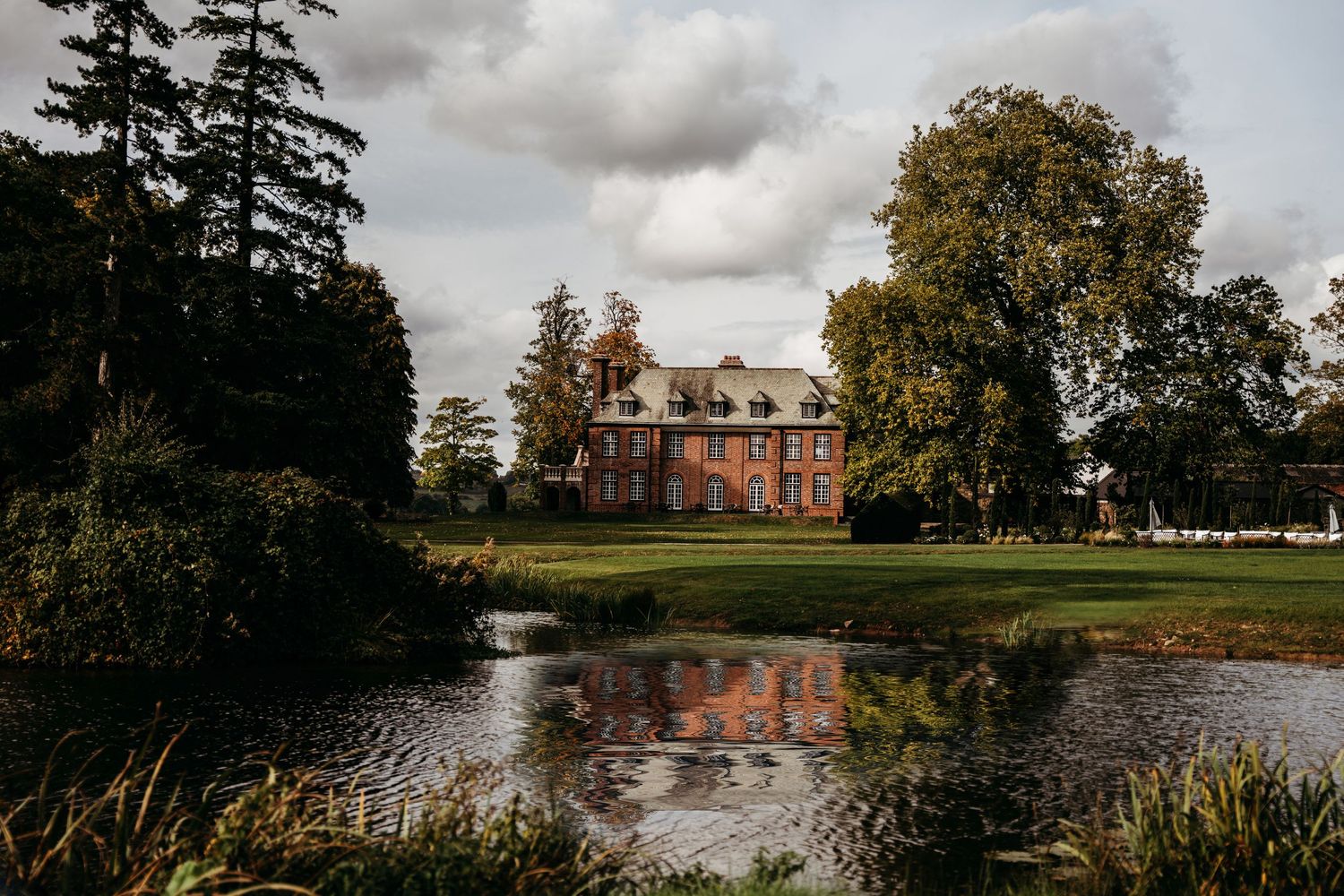 Grand estate house reflected in pond with dramatic cloudy sky and autumn trees.