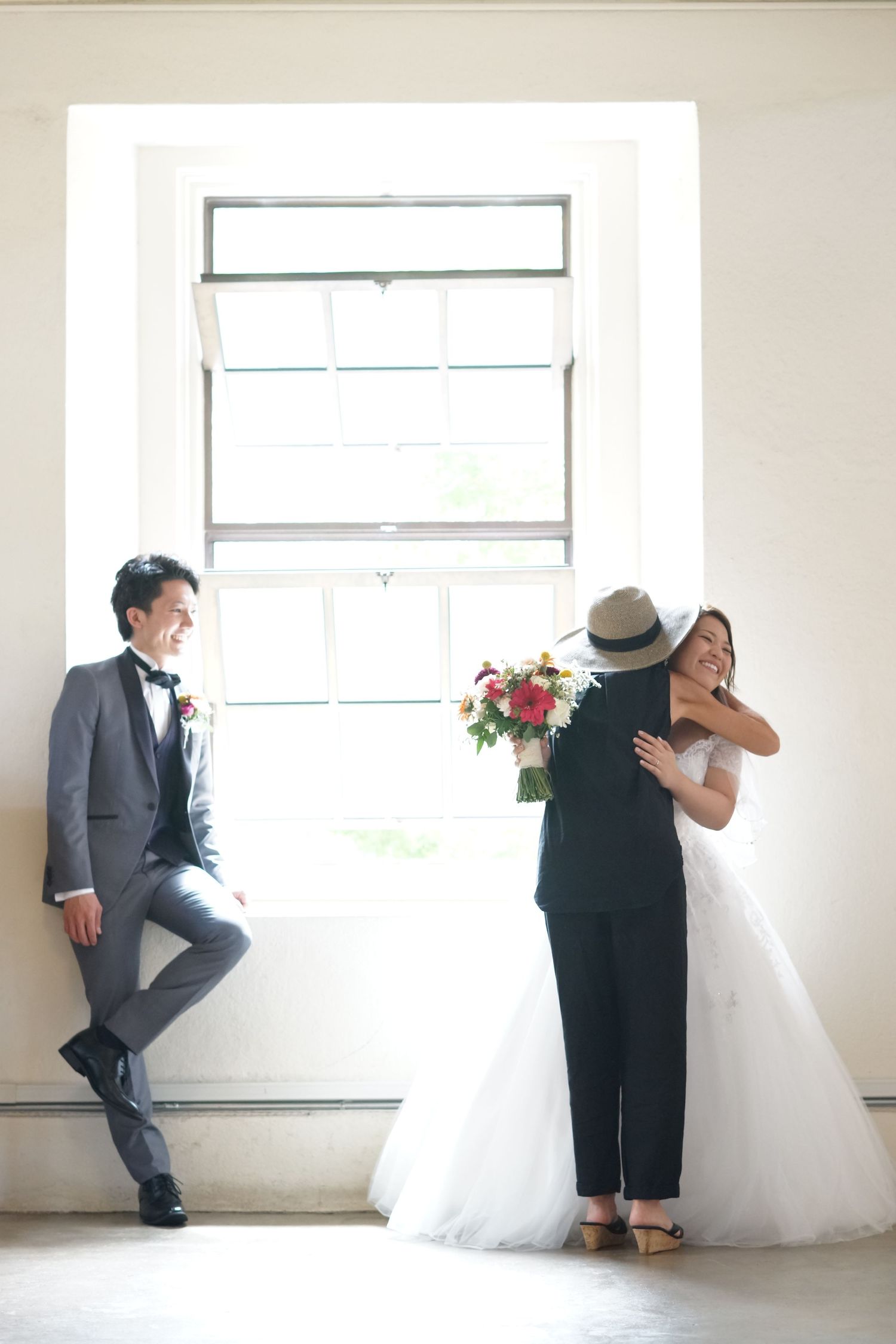 Groom in gray tuxedo watches bride receive congratulatory hug by bright window indoors.
ハワイウェディング
ハワイ挙式